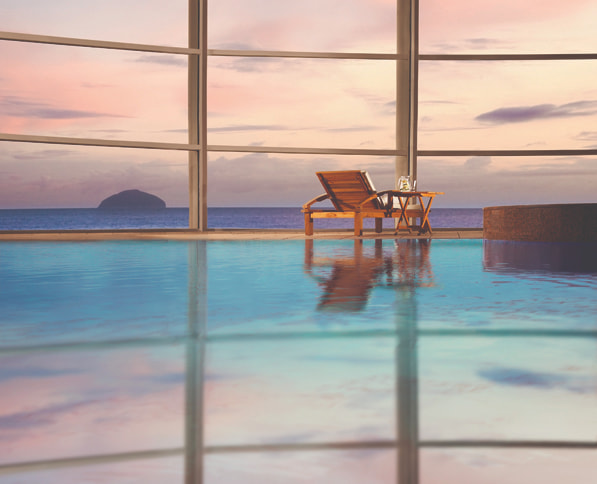 Calm indoor pool with reclined seats, looking out through a glass wall onto the ocean.