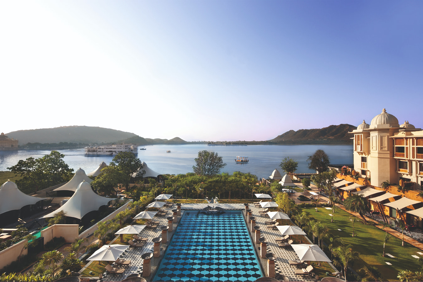 Arial view of an outdoor pool, with shaded reclined beds and a nearby grassy area, looking out onto the ocean.