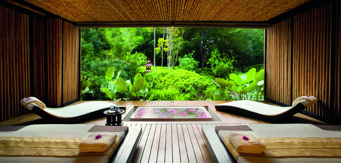 Wooden-walled room with a pool and reclined beds, looking out through an open wall onto lush vegetation.